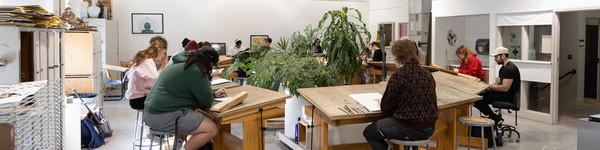 Art students drawing at large wooden desks set in circle around plants.