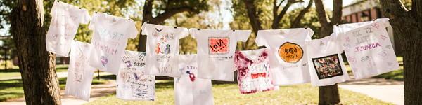 On a clothes line between two trees, hangs painted t-shirts about surviving sexual assault.